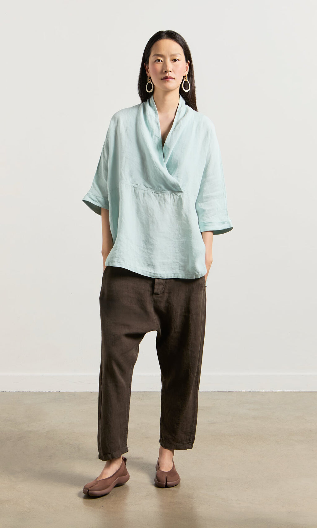 Plümo- Woman in a wrapover blouse with cropped sleeves, loose brown pants, and brown flats against a plain light background.