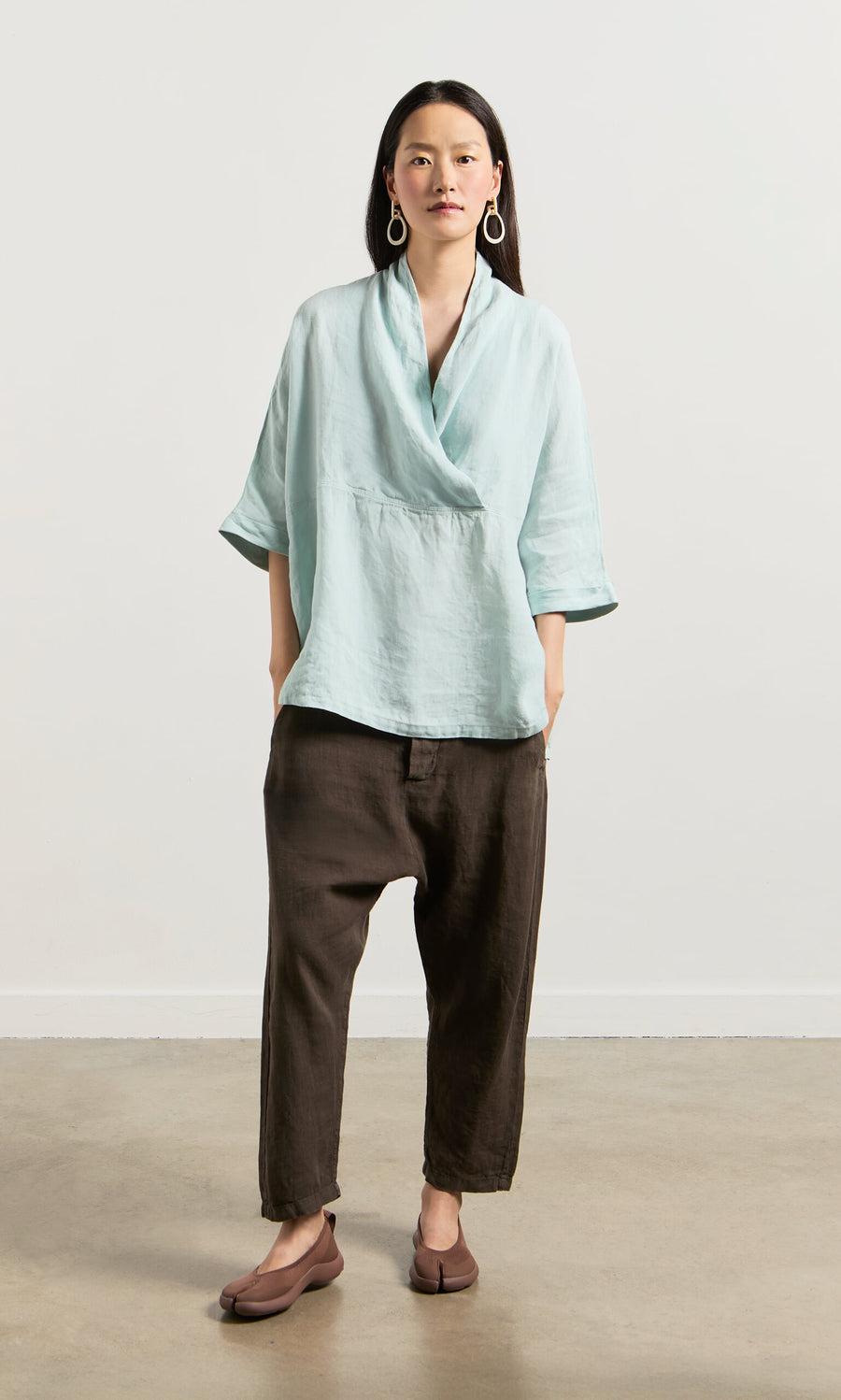 Plümo- Woman in a wrapover blouse with cropped sleeves, loose brown pants, and brown flats against a plain light background.