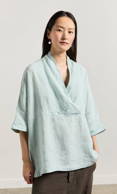 Plümo- A woman in a Wrapover Blouse with cropped sleeves and dark pants stands facing the camera against a light background.