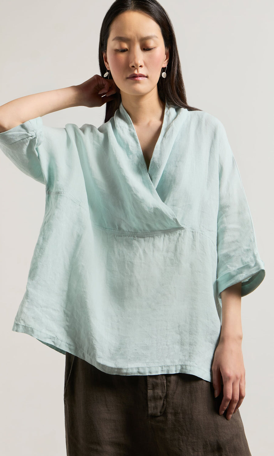 Plümo- Woman in a loose, light blue Wrapover Blouse with cropped sleeves, posing with one hand behind her neck.
