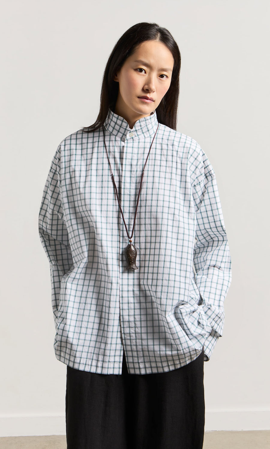 Plümo- Woman in a checked shirt and black pants wears the Yumi Fish Necklace, standing with hands in pockets.