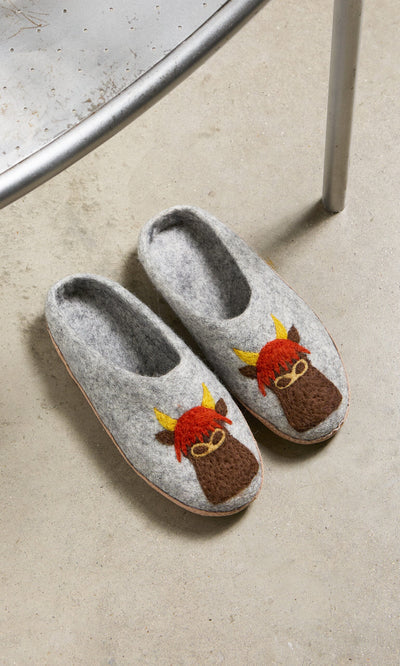 Plümo- Highland Slippers—gray cow design with yellow horns and red hair—on a concrete floor by a metal chair.