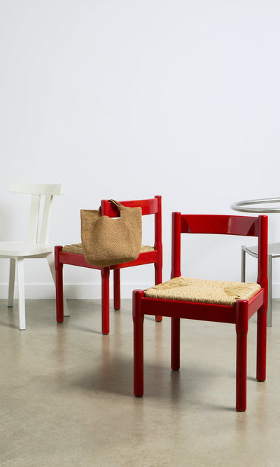 Plümo- A red chair with a Tara Straw Bag sits beside another red chair, a white chair, and a round table on concrete.