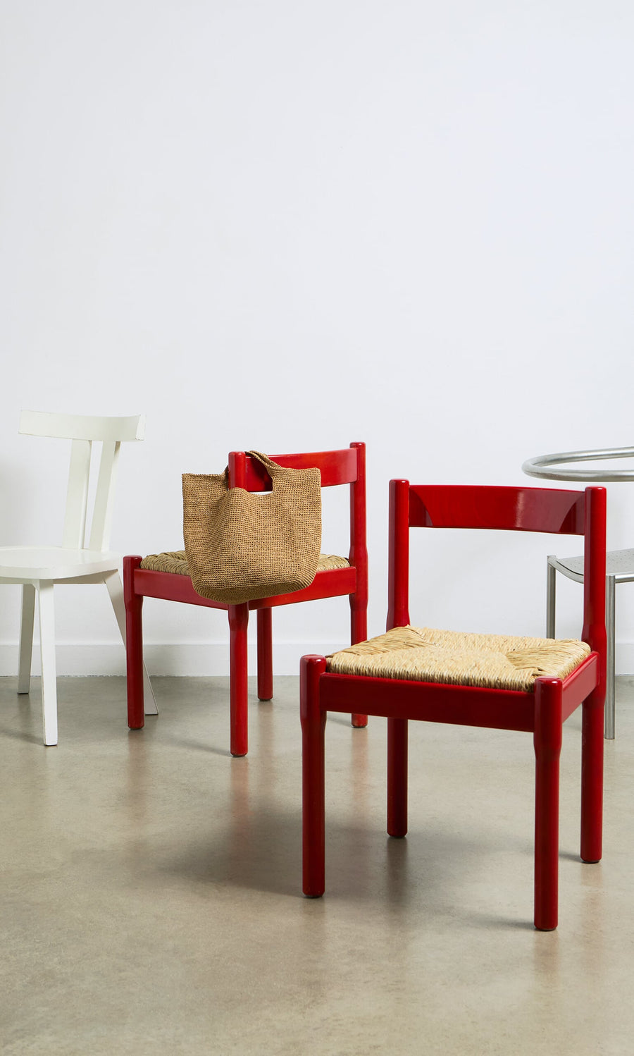 Plümo- A red chair with a Tara Straw Bag sits beside another red chair, a white chair, and a round table on concrete.