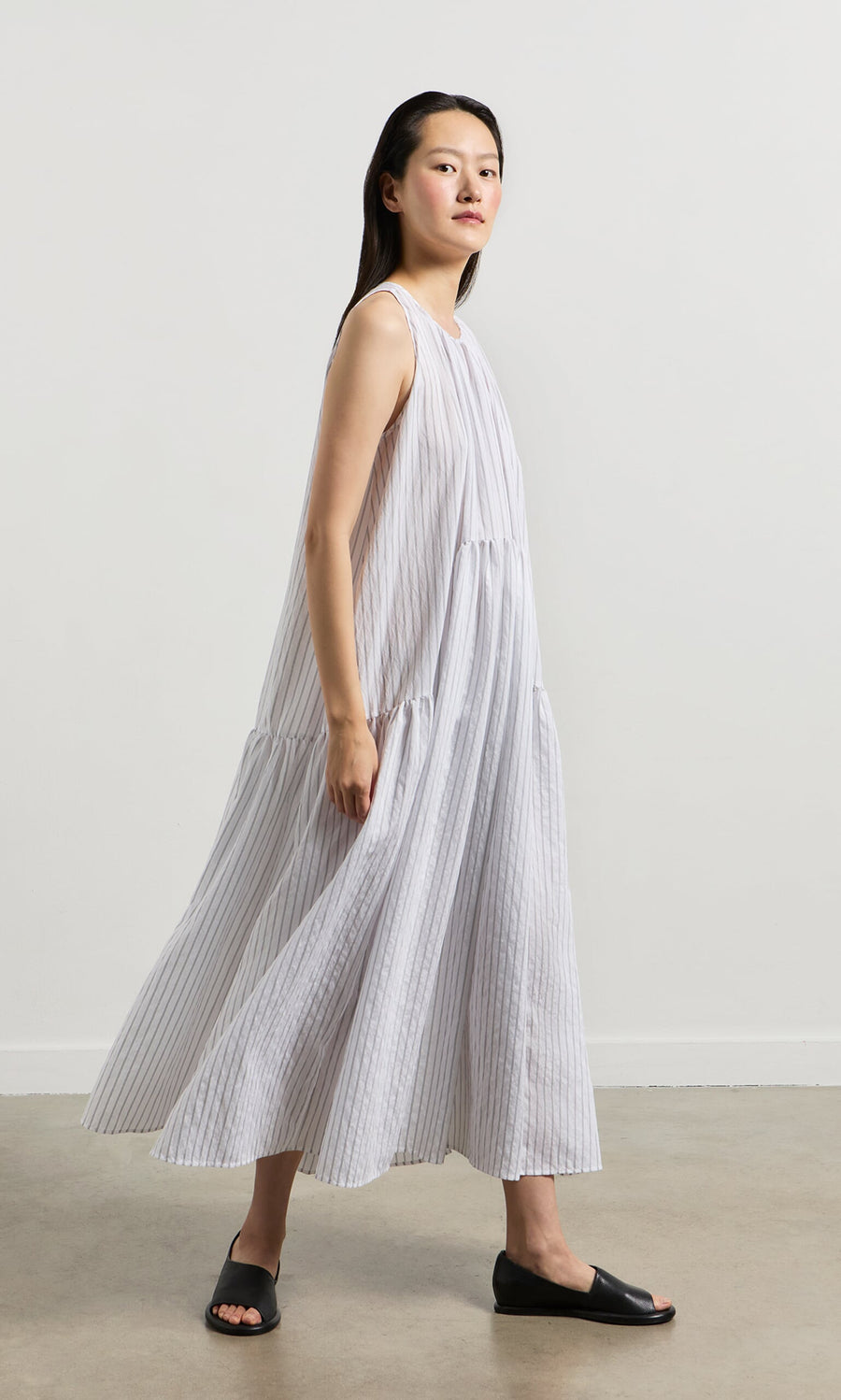 Plümo- Woman wearing the Alba Stripe Dress and black sandals walking indoors against a plain background.