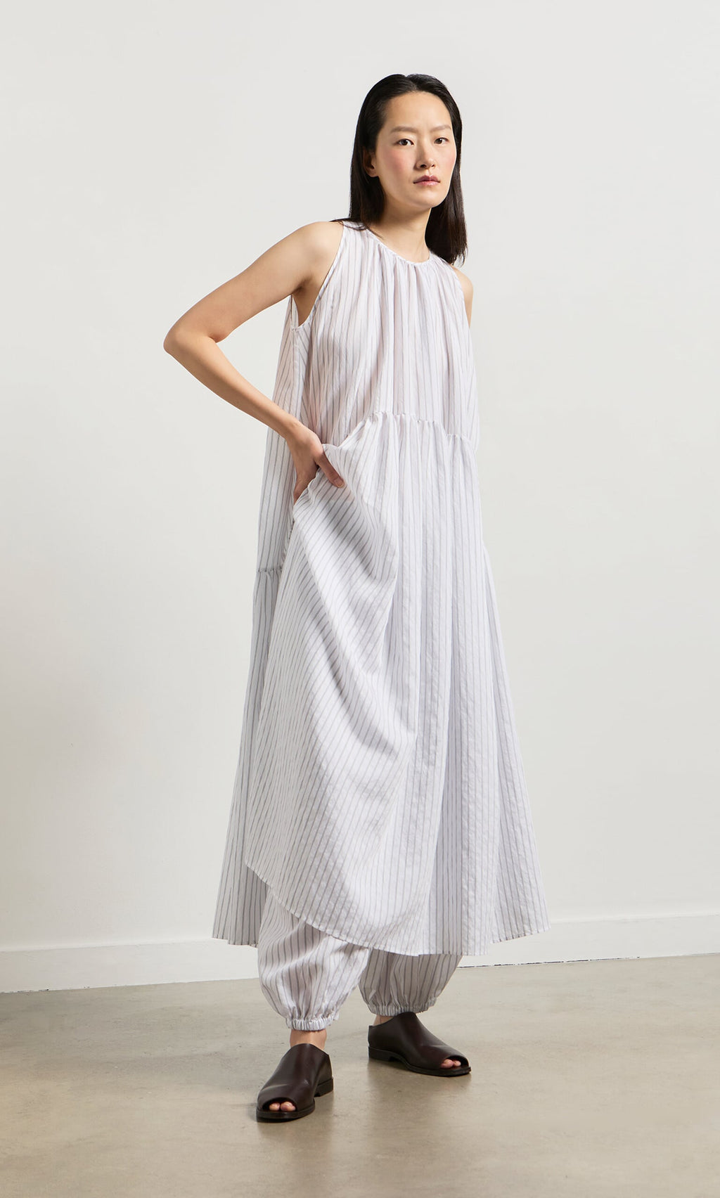 Plümo- Woman wearing the Alba Stripe Dress, a loose white pinstripe dress over matching pants, on a neutral background.