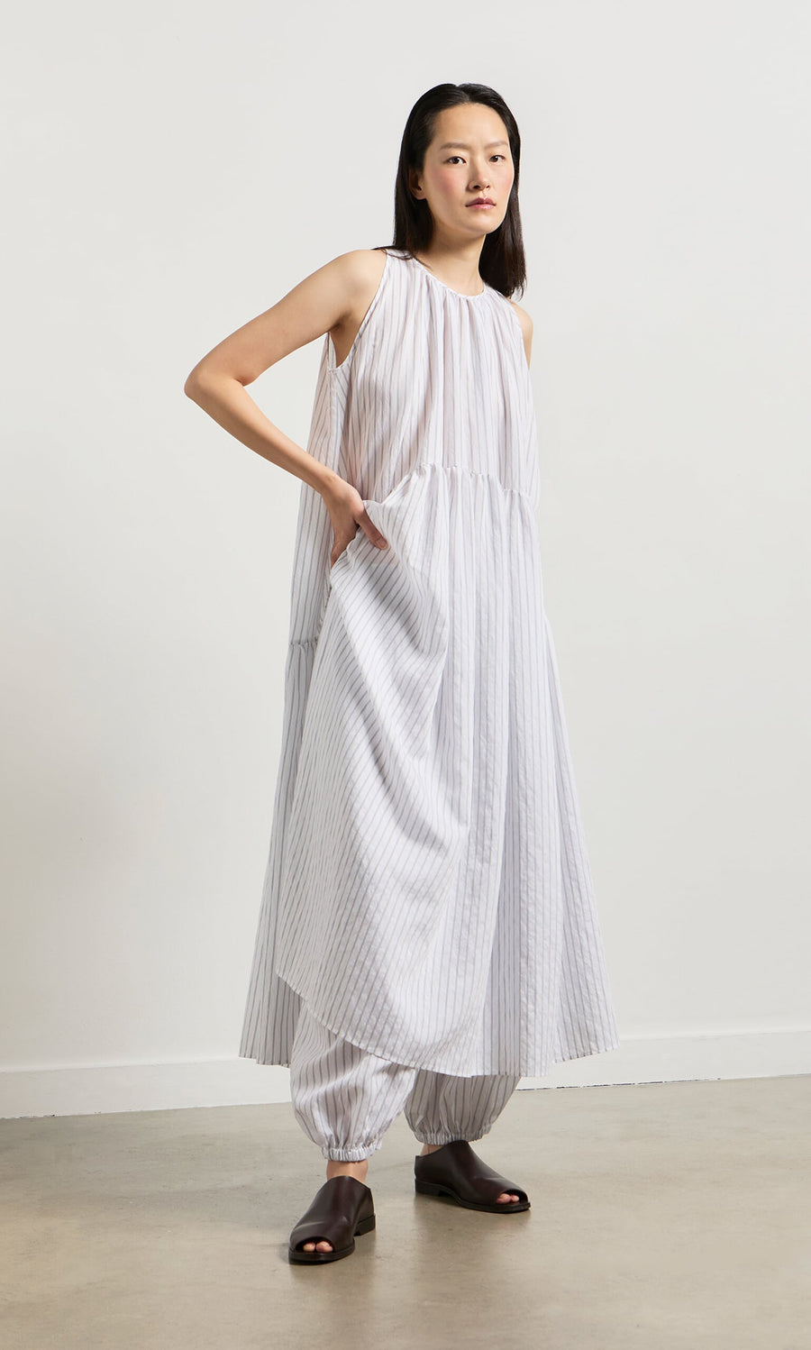 Plümo- Woman wearing the Alba Stripe Dress, a loose white pinstripe dress over matching pants, on a neutral background.