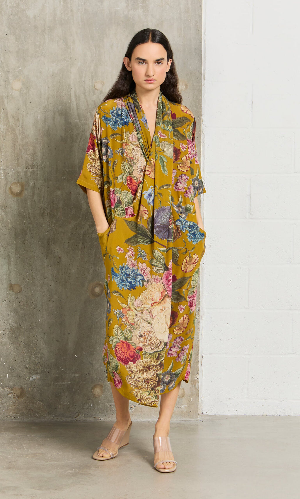 Plümo- Someone wears the Amber Twist Dress, a loose mustard-yellow botanical print, by a concrete and white brick wall.