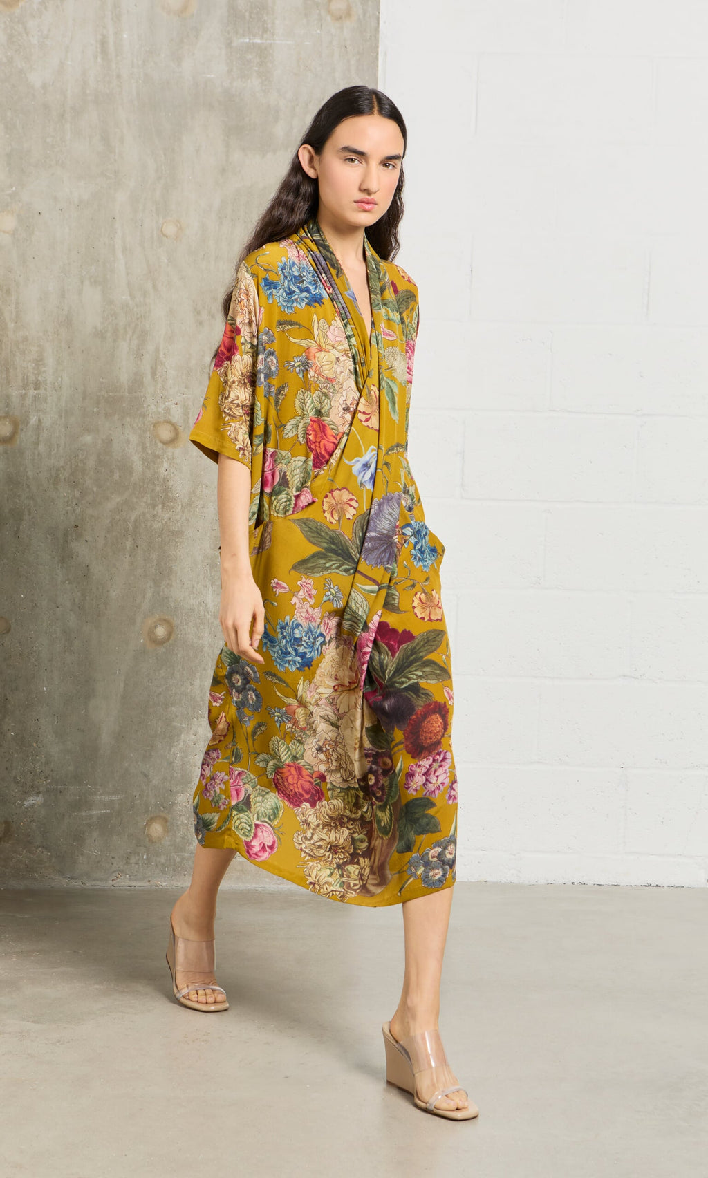 Plümo- Woman in the Amber Twist Dress and nude wedge sandals walks indoors against a concrete and white wall background.