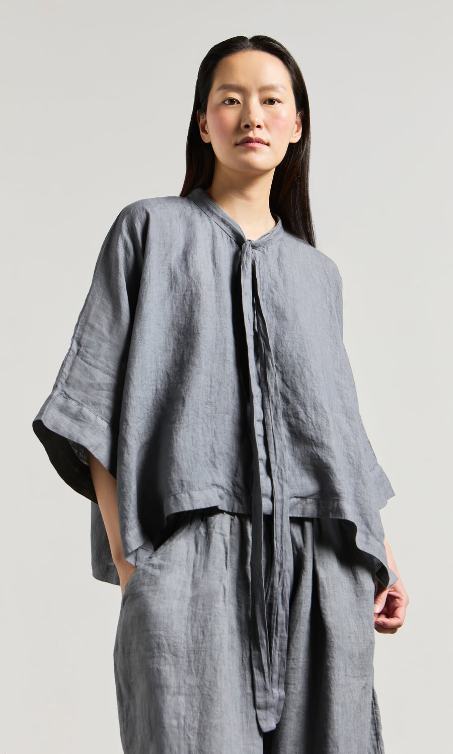 Plümo- Woman in the Antoine Blouse - Grey, a loose linen top with ribbon necktie, made in Greece.