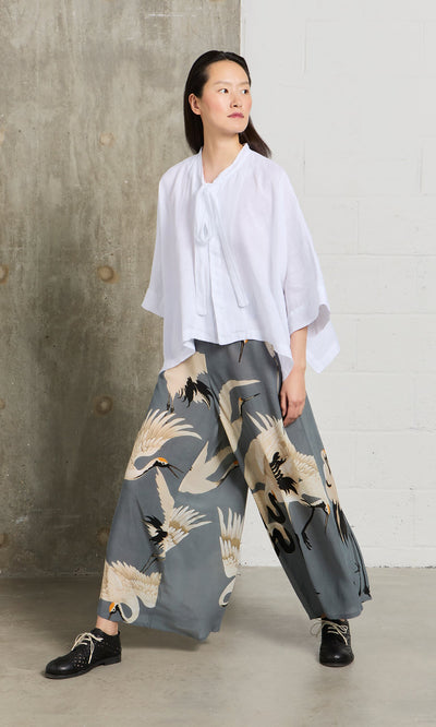 Plümo- A woman in a white blouse and Slate Stork Pants stands by a concrete and white wall.