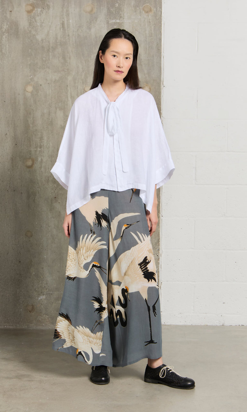 Plümo- Woman in a white blouse and Slate Stork Pants stands indoors by a white and concrete wall.