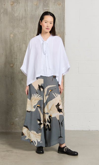 Plümo- Woman in a white blouse and Slate Stork Pants stands indoors by a white and concrete wall.