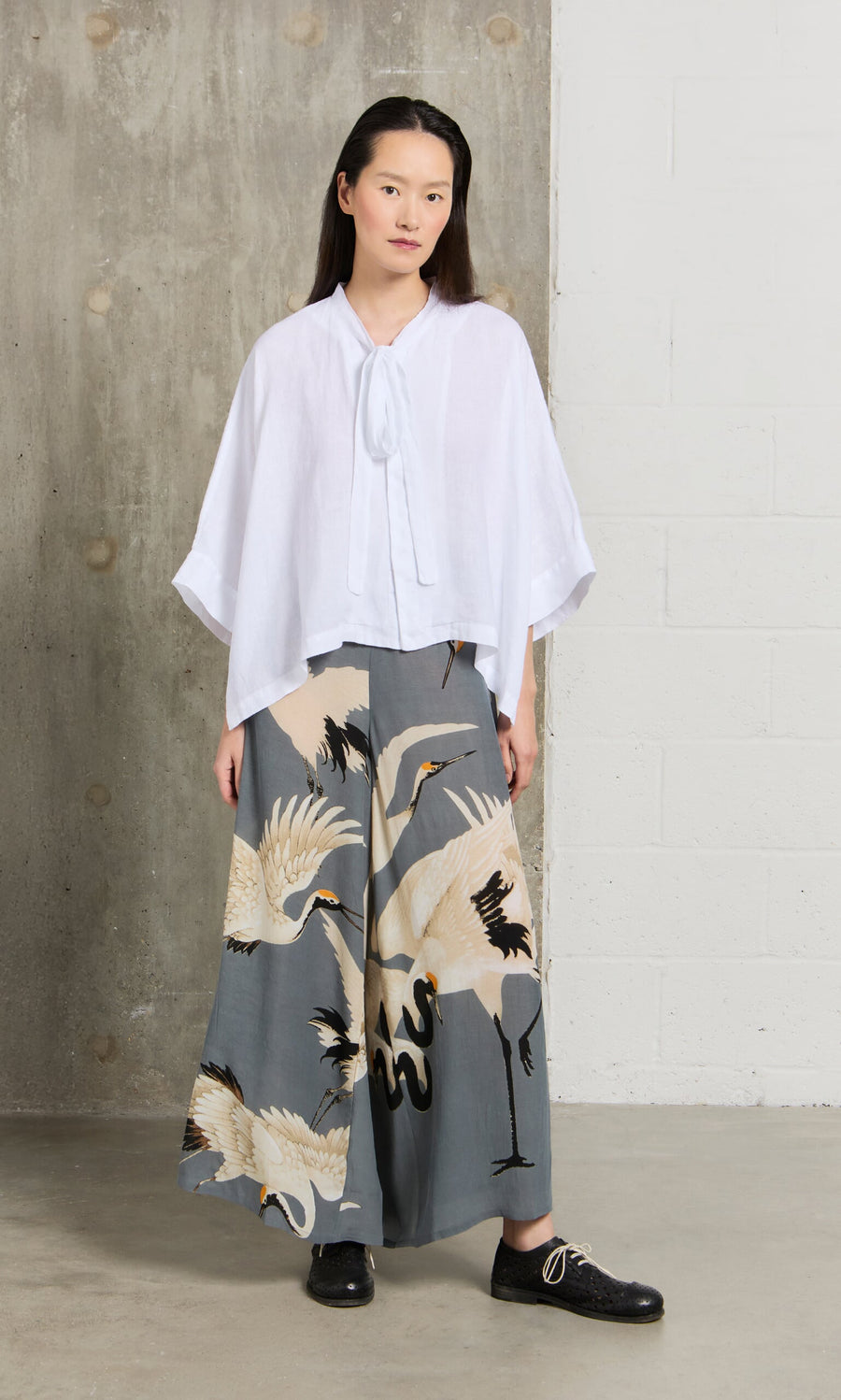 Plümo- Woman in a white blouse and Slate Stork Pants stands indoors by a white and concrete wall.