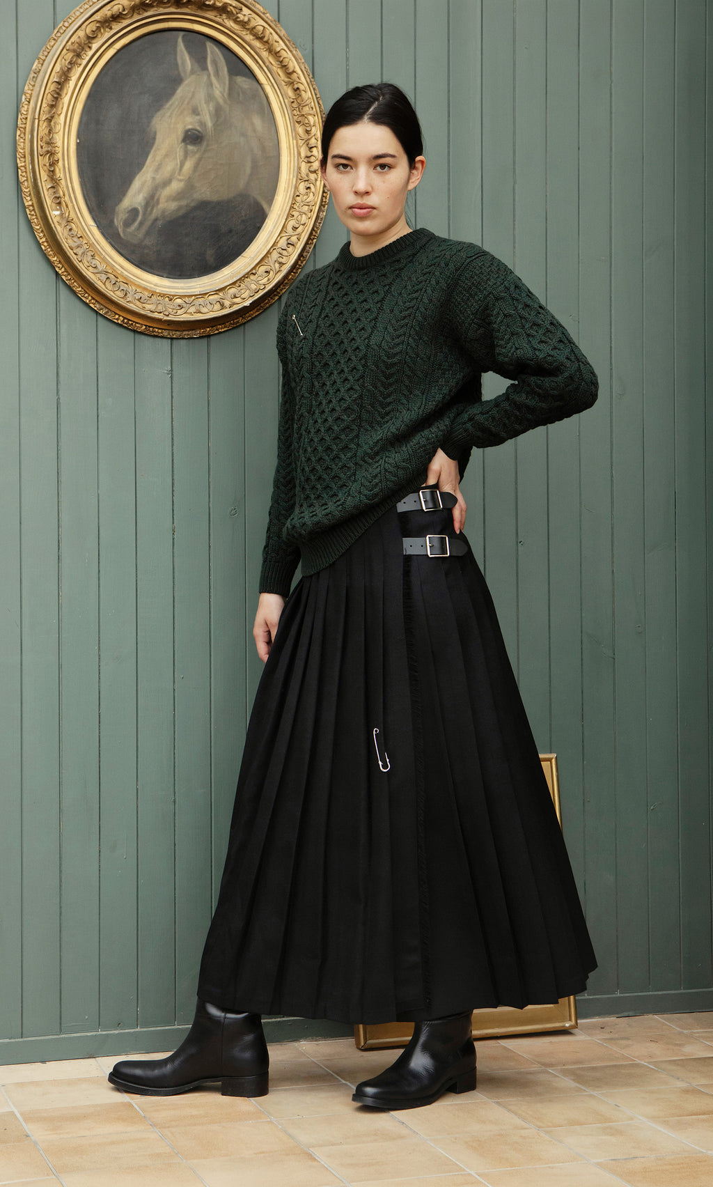 Plümo  Woman in a Croft Green Aran Jumper and black skirt stands indoors by a horse painting on a green wooden wall.