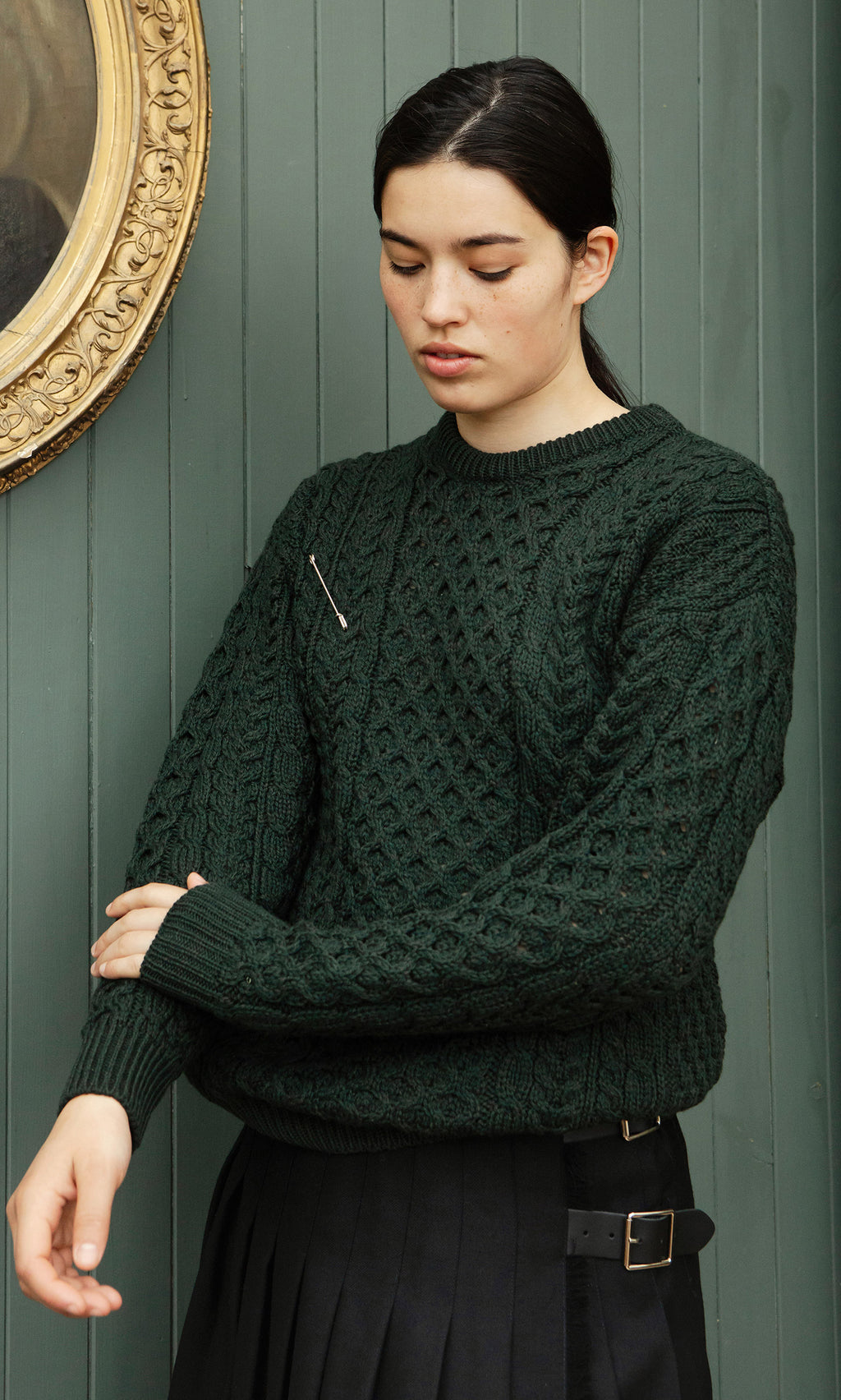 Plümo  Person wears Croft Green Aran Jumper and black skirt, standing against green paneled wall, looking down.