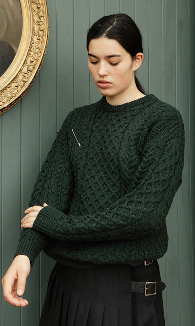Plümo  Person wears Croft Green Aran Jumper and black skirt, standing against green paneled wall, looking down.