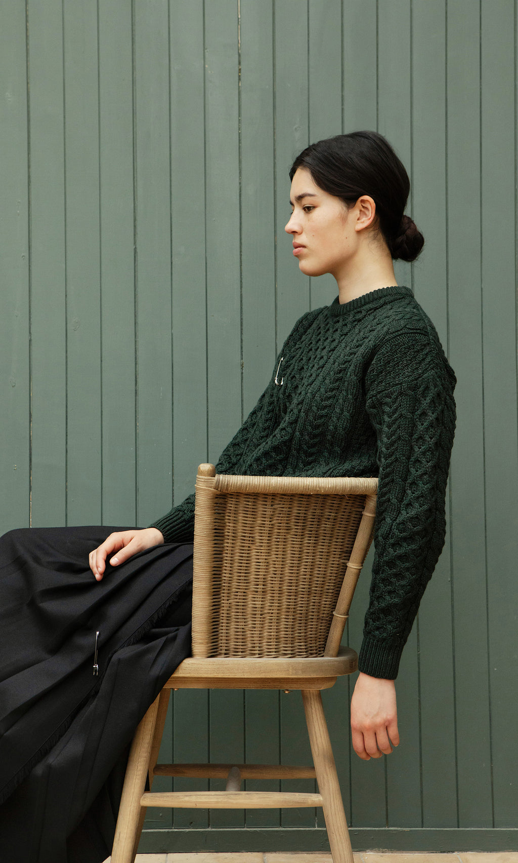 Plümo  Woman in an Aran Jumper - Croft Green and black skirt sits sideways on a chair by a green paneled wall.