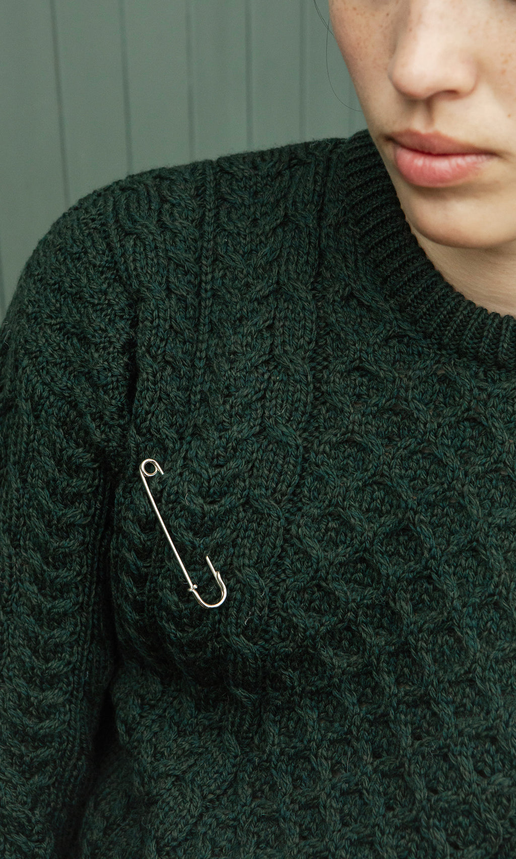Plümo  Individual in a Croft Green Aran Jumper with a safety pin on the shoulder, turned partially away.