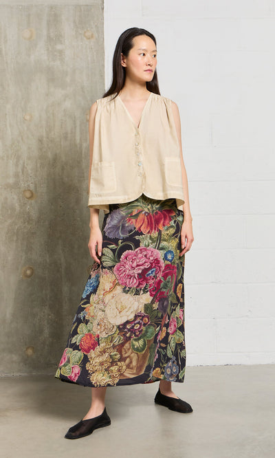 Plümo- A woman in a beige sleeveless top and the Auricula Skirt stands indoors against a concrete and brick wall.