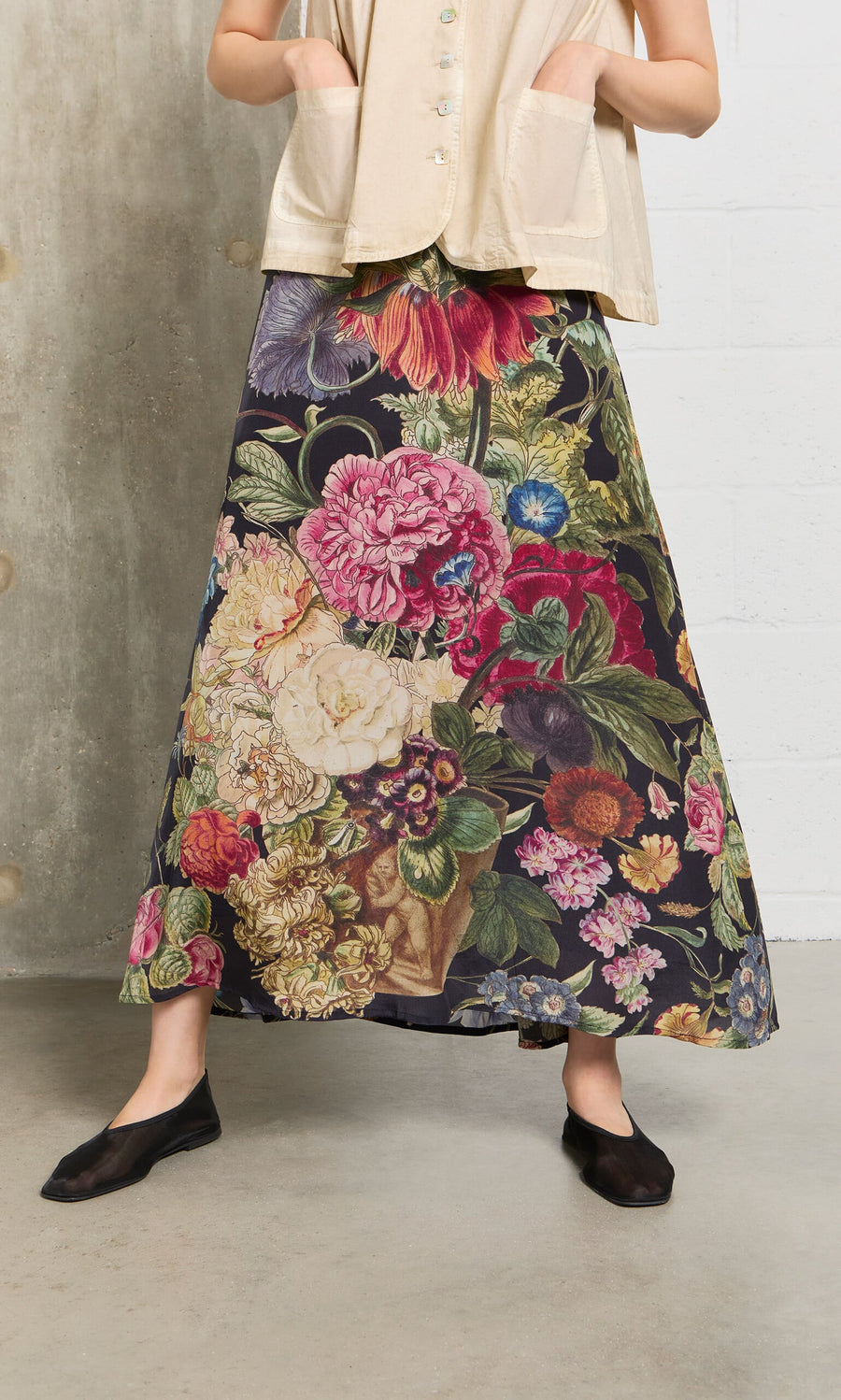 Plümo- Woman in a beige top and the Auricula Skirt, an A-line black botanical print, standing indoors.