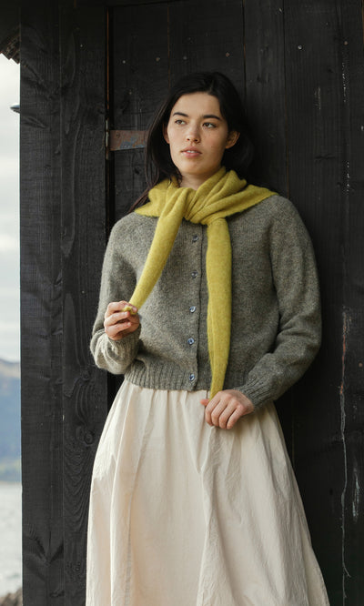 Plümo  Woman in a grey sweater and Avo Necktie stands by a dark wood door, holding something and looking ahead.