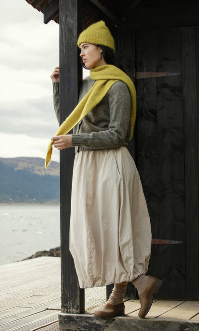 Plümo  Woman in an Avo Wool Beanie, yellow scarf, and cream skirt stands by a dark wooden structure near water.