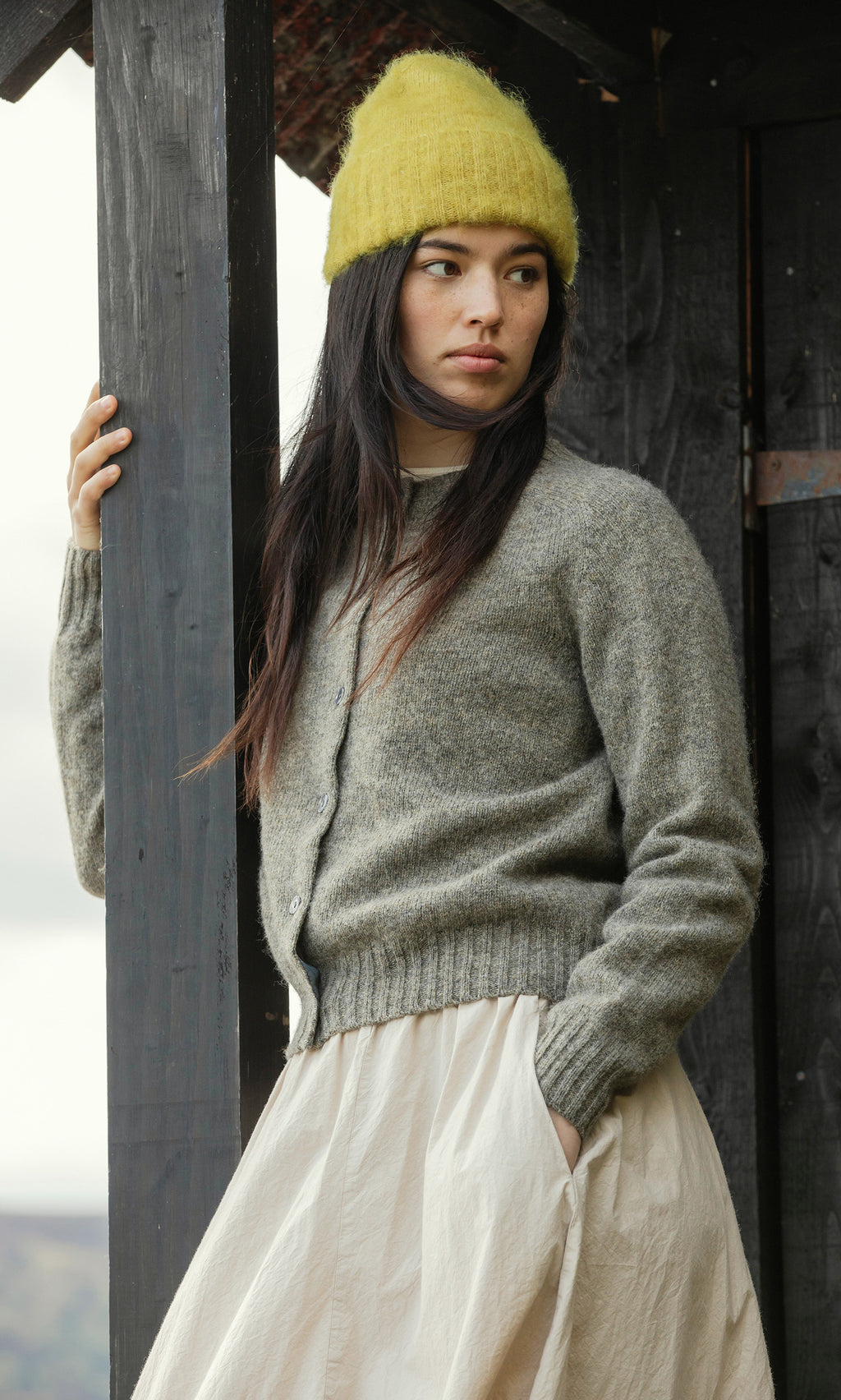 Plümo  Woman in Avo Wool Beanie, gray cardigan, and cream skirt stands by a wooden post, looking to the side.
