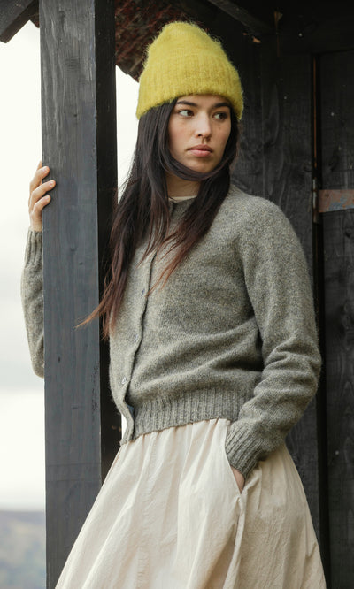 Plümo  Woman in Avo Wool Beanie, gray cardigan, and cream skirt stands by a wooden post, looking to the side.