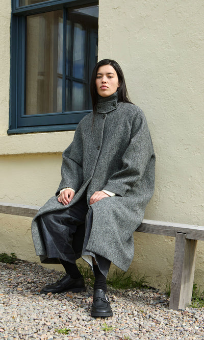 Plümo  A person sits on a wooden bench wearing a grey herringbone Balmacaan Tweed Coat with a high collar and dark shoes.