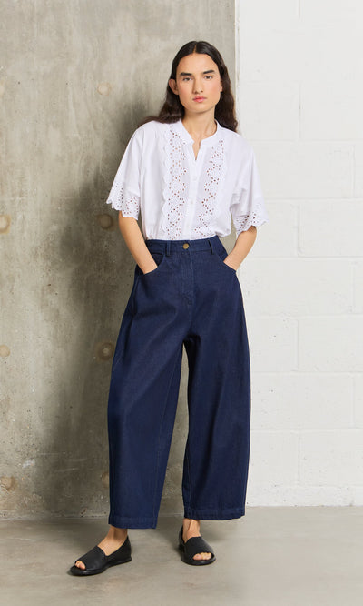 Plümo- Woman in Barrel Pants Indigo, a white eyelet blouse, and black sandals stands against a concrete wall.