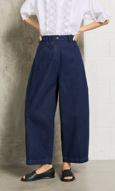 Plümo- Person wears Barrel Pants Indigo, a white eyelet blouse, and black open-toe flats against a concrete wall.