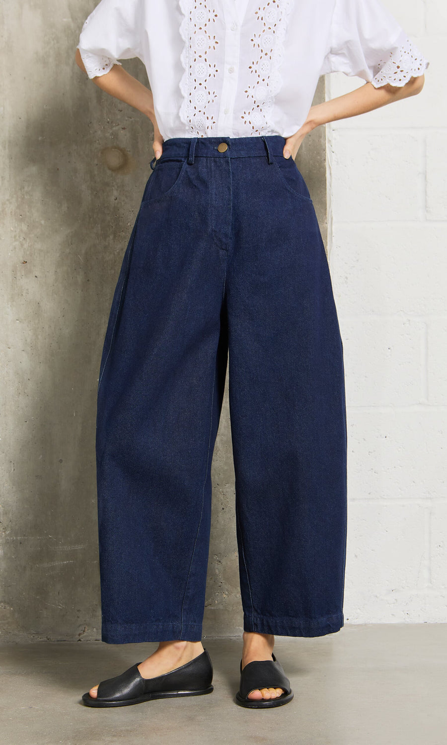 Plümo- Person wears Barrel Pants Indigo, a white eyelet blouse, and black open-toe flats against a concrete wall.