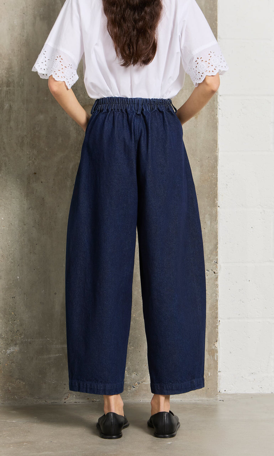 Plümo- Person standing with back to camera, wearing Barrel Pants Indigo jeans, a white blouse, and black shoes.