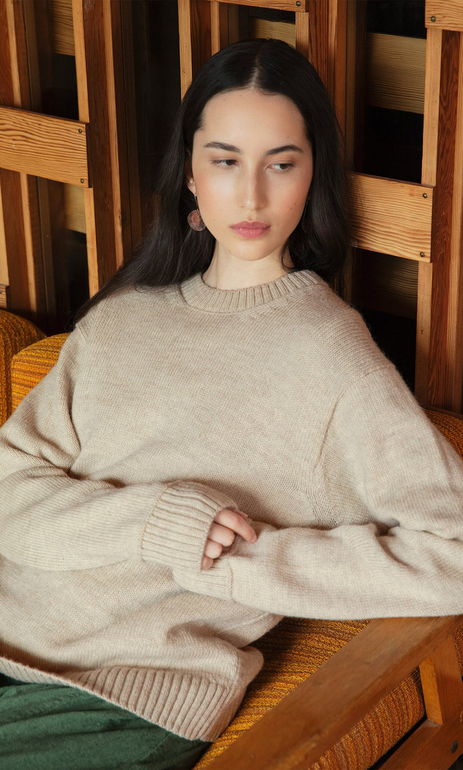 Plümo- Woman with long dark hair in the Christie Wool Jumper sits on a wooden chair, looking to the side calmly.