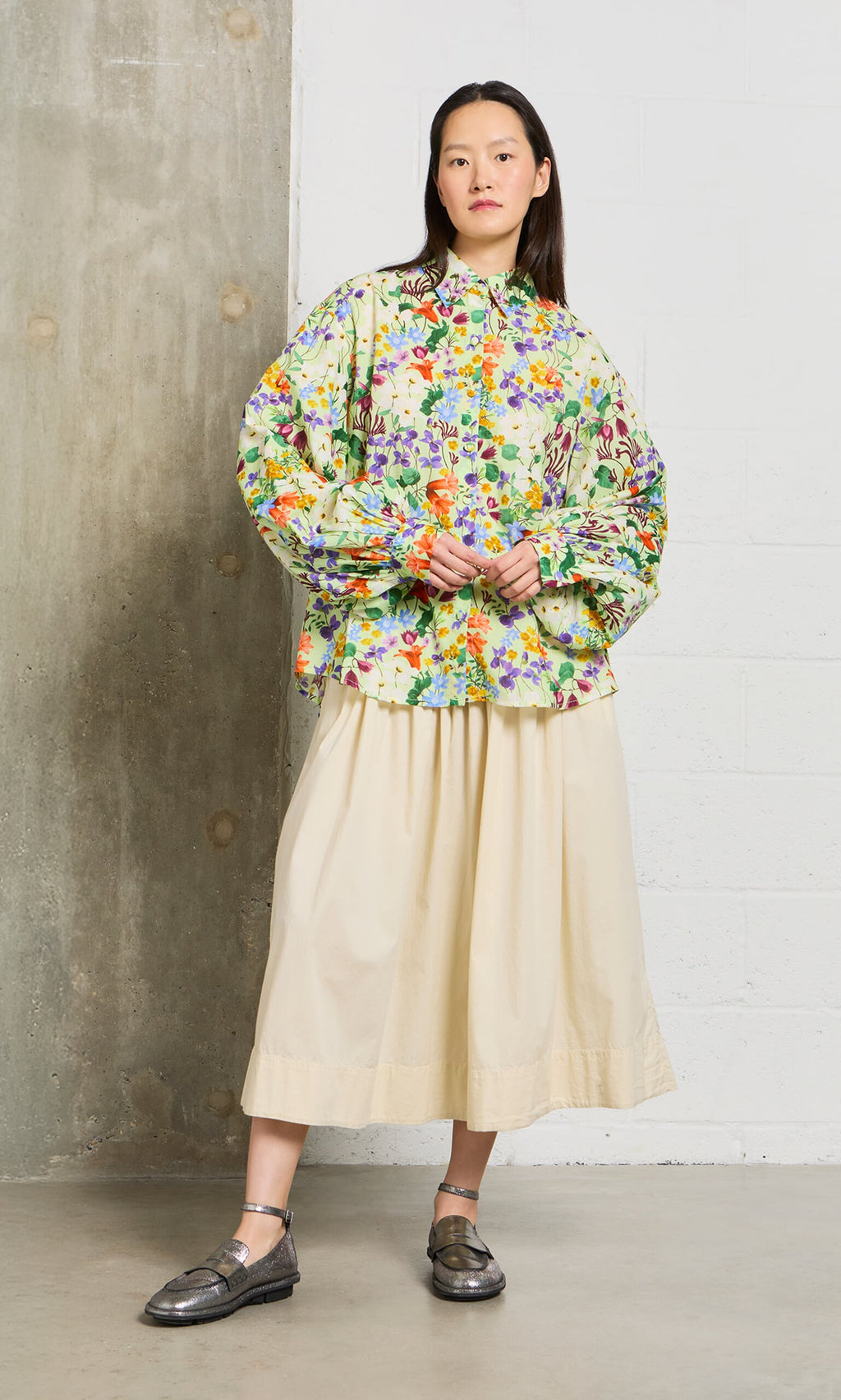 Plümo- A woman in a floral blouse and the Rika Wide Skirt with slip pockets stands by a concrete and white brick wall.