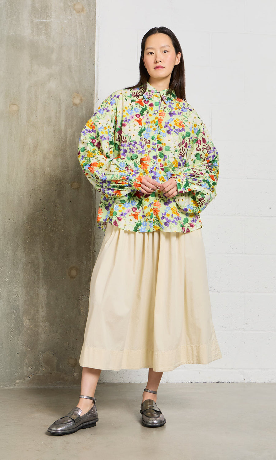 Plümo- A woman in a floral blouse and the Rika Wide Skirt with slip pockets stands by a concrete and white brick wall.