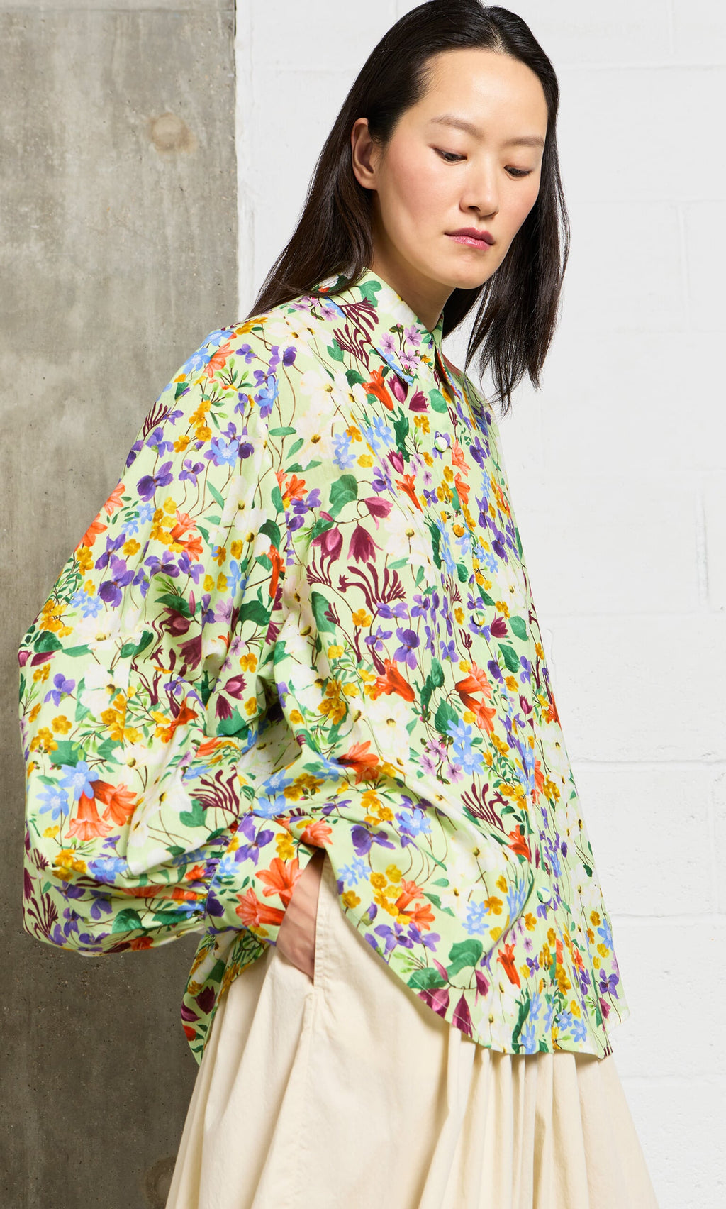 Plümo- Woman wears the Beauvais Blouse, a jewel-toned botanical shirt with balloon sleeves, against a concrete wall.