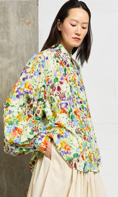 Plümo- Woman wears the Beauvais Blouse, a jewel-toned botanical shirt with balloon sleeves, against a concrete wall.