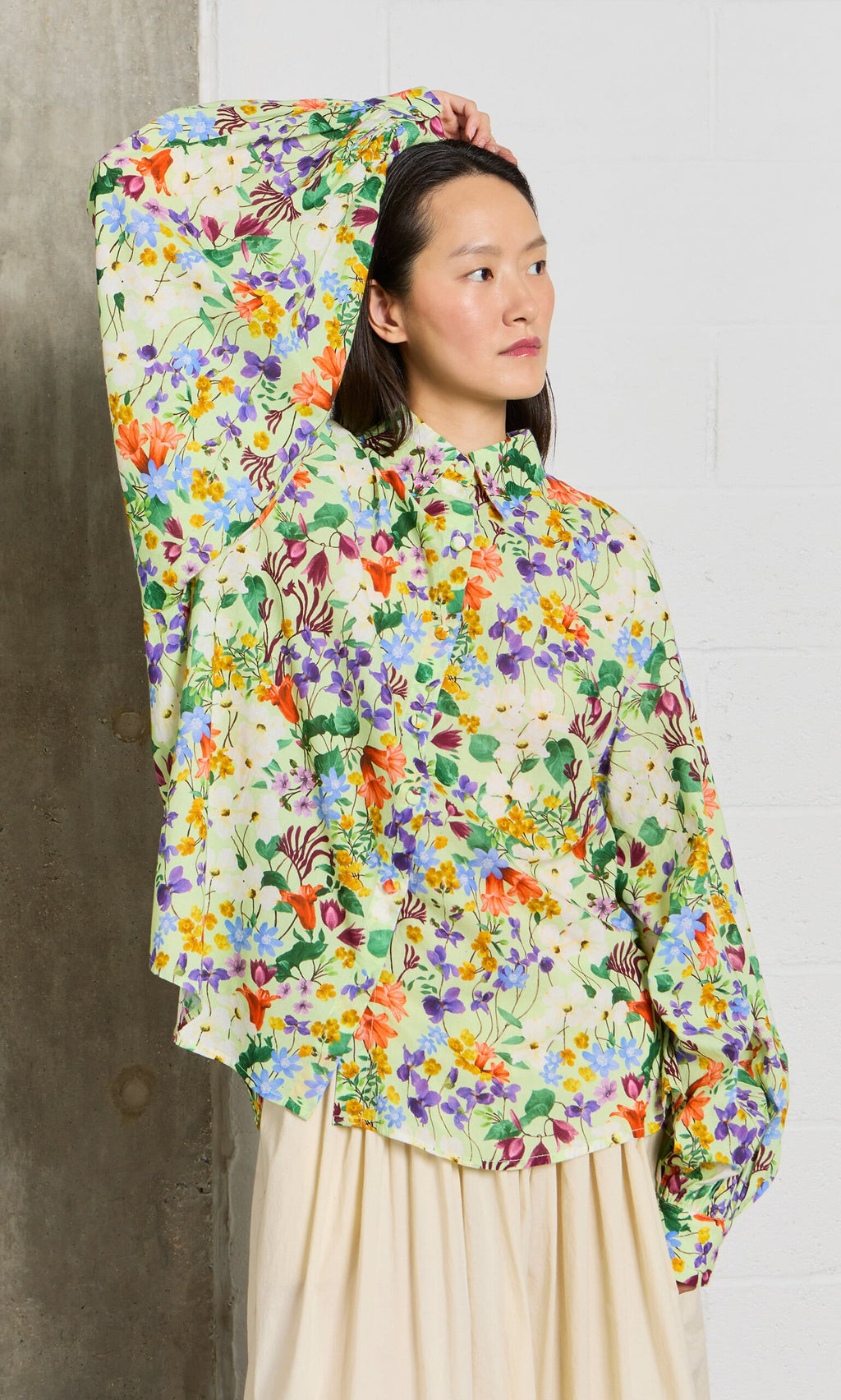 Plümo- Model wears the Beauvais Blouse in jewel-toned botanical print, posing with one sleeve raised before a white brick wall.