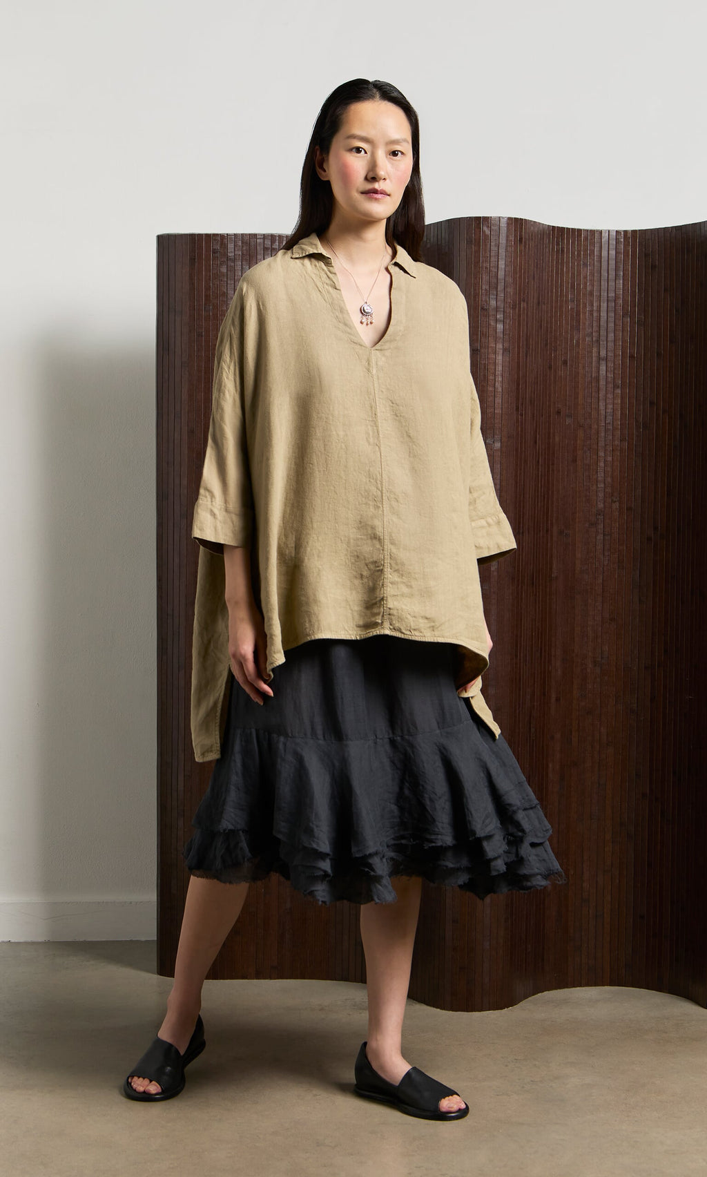 Plümo- Woman in a Bergman Blouse, black ruffled skirt, and sandals stands indoors by a brown folding screen.