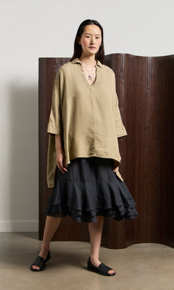 Plümo- Woman in a Bergman Blouse, black ruffled skirt, and sandals stands indoors by a brown folding screen.