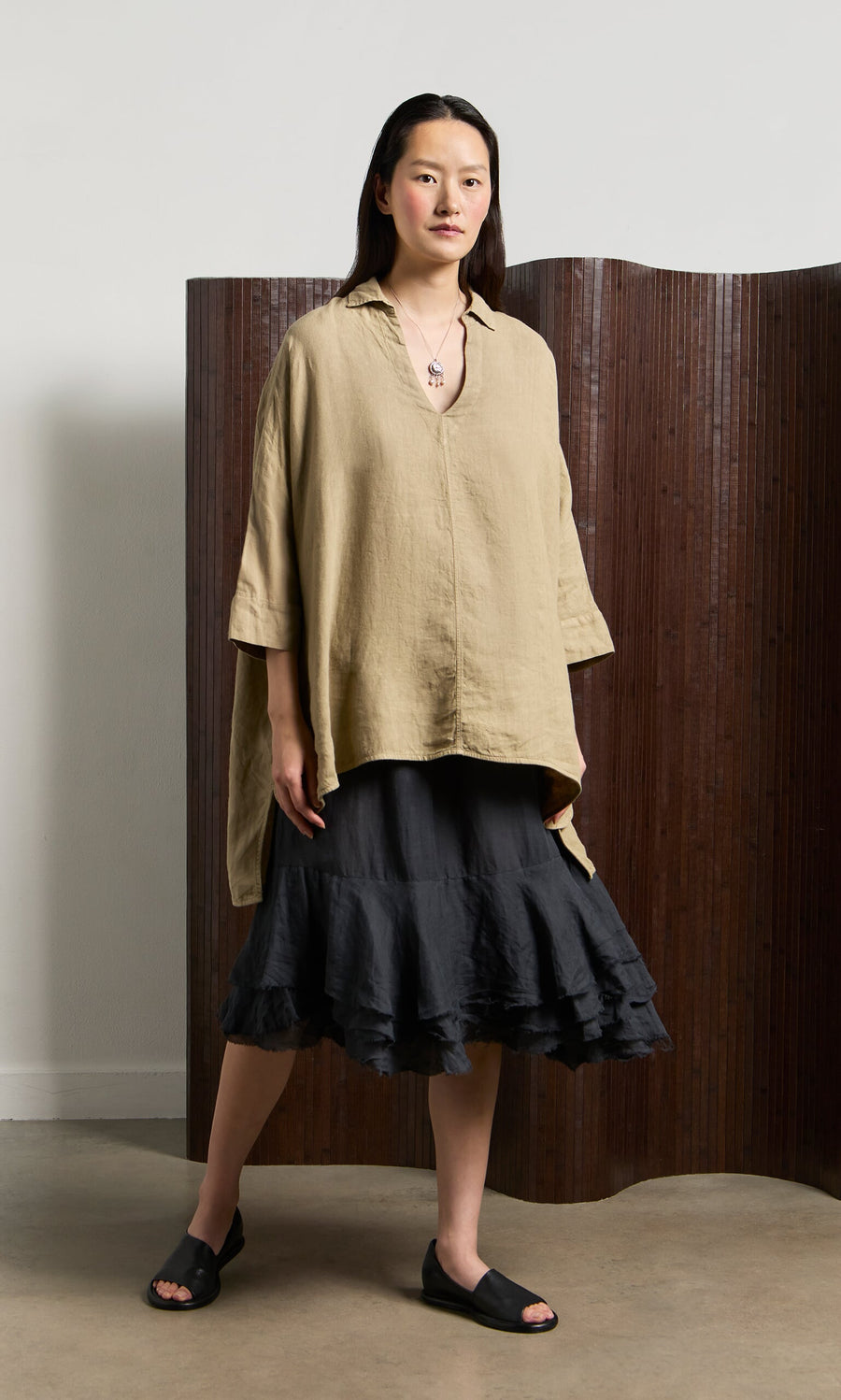 Plümo- Woman in a Bergman Blouse, black ruffled skirt, and sandals stands indoors by a brown folding screen.