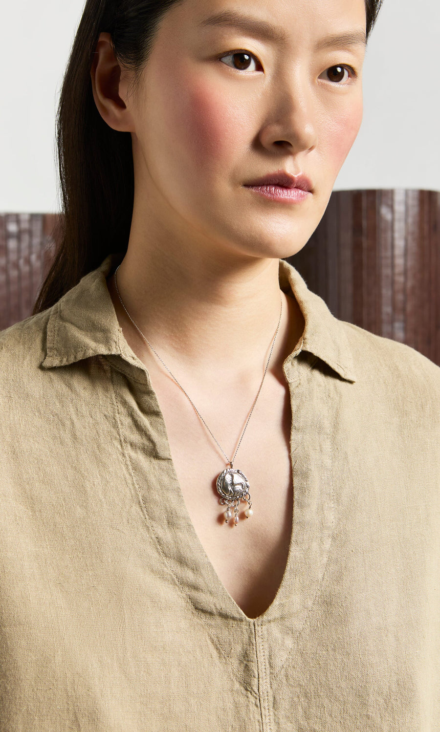 Plümo- Woman wearing the Bergman Blouse, a garment-dyed linen V-neck top, paired with a silver necklace indoors.
