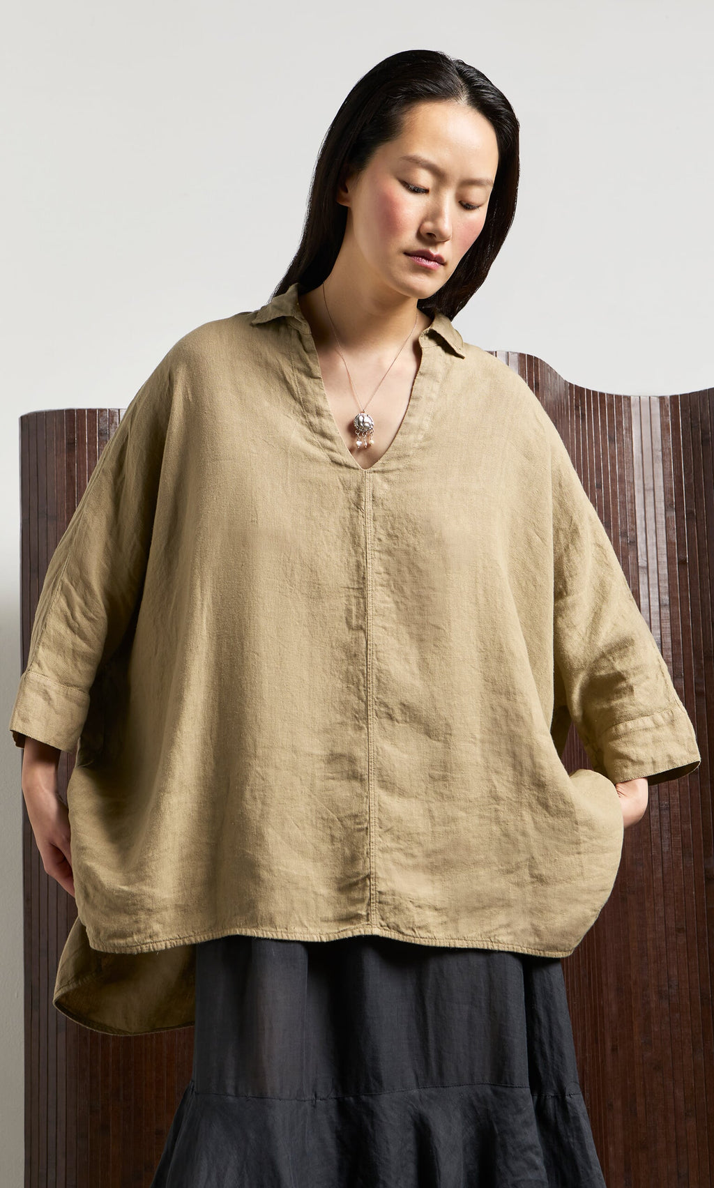 Plümo- Woman in an oversized V-neck Bergman Blouse and black skirt stands against a wooden screen, looking down.