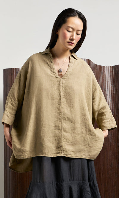 Plümo- Woman in an oversized V-neck Bergman Blouse and black skirt stands against a wooden screen, looking down.