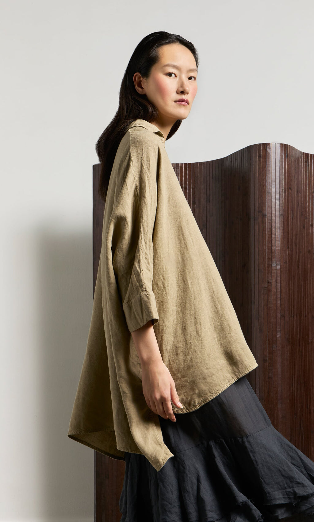 Plümo- Woman in a Bergman Blouse and black skirt stands by a wooden room divider, looking to the side.