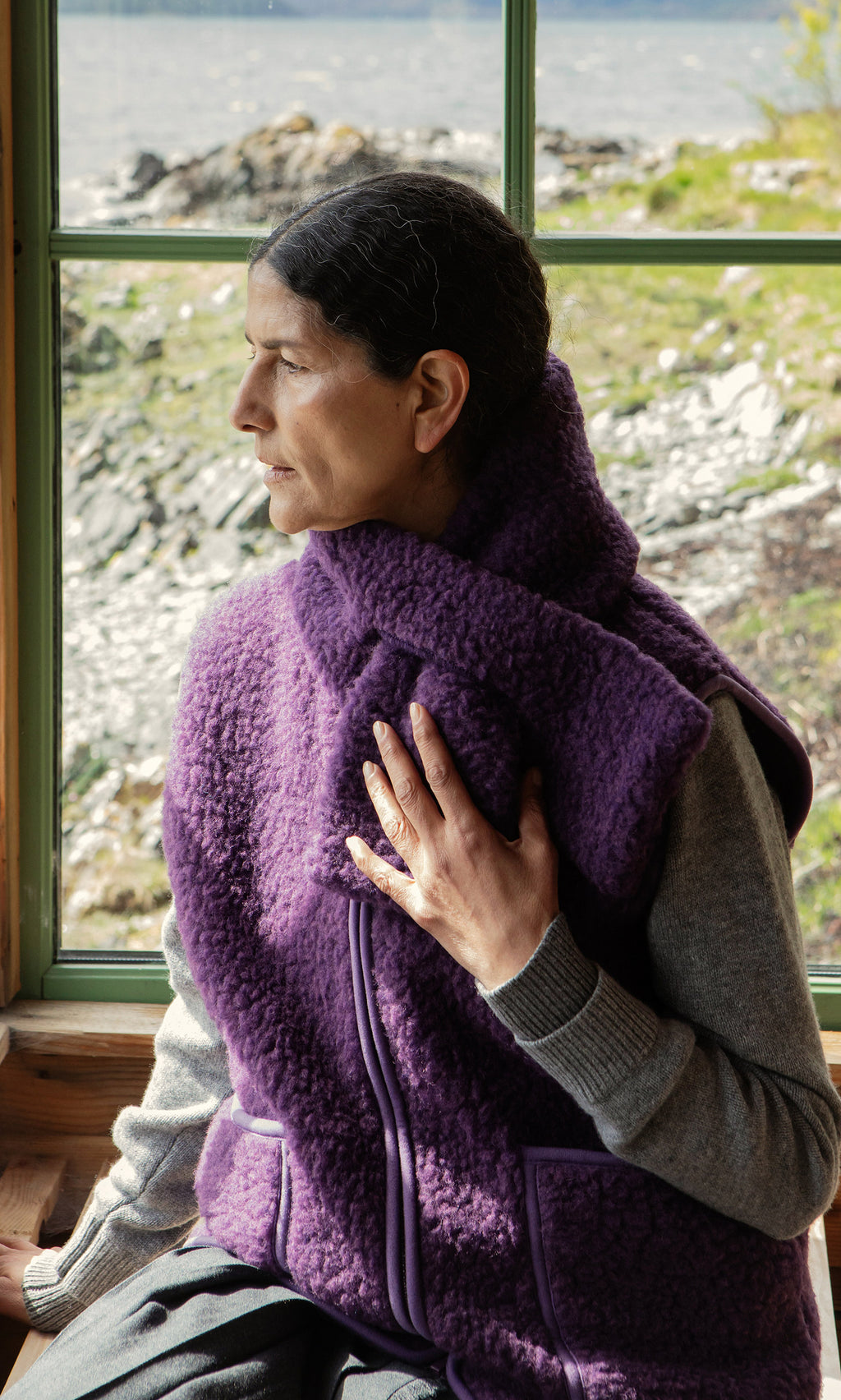 Plümo  Woman in a purple fleece vest wears the Betty Scarf as she sits by a window, gazing thoughtfully outside.