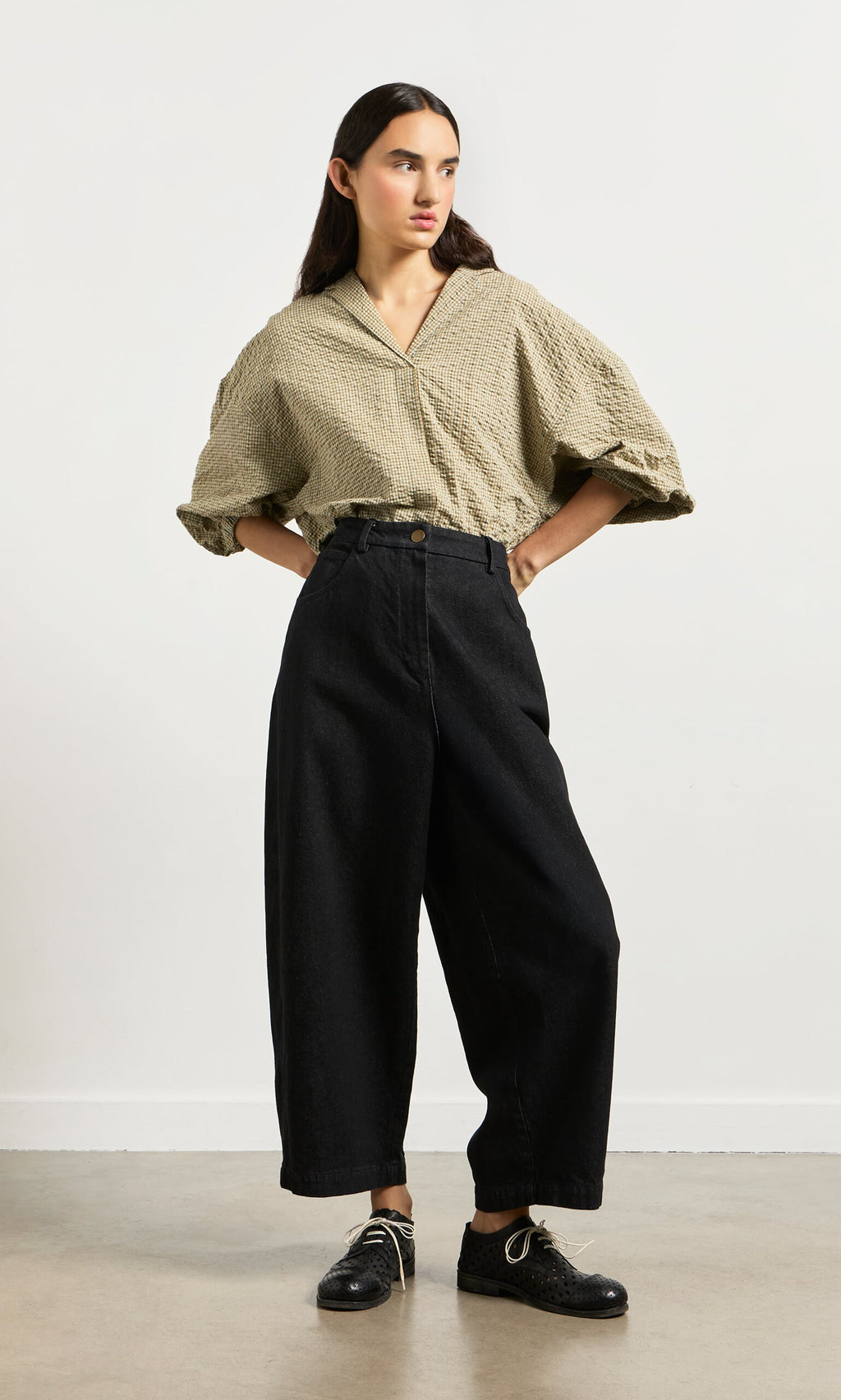 Plümo- Woman stands against a plain wall wearing loose Black Barrel Pants and a beige blouse, embracing a relaxed silhouette.