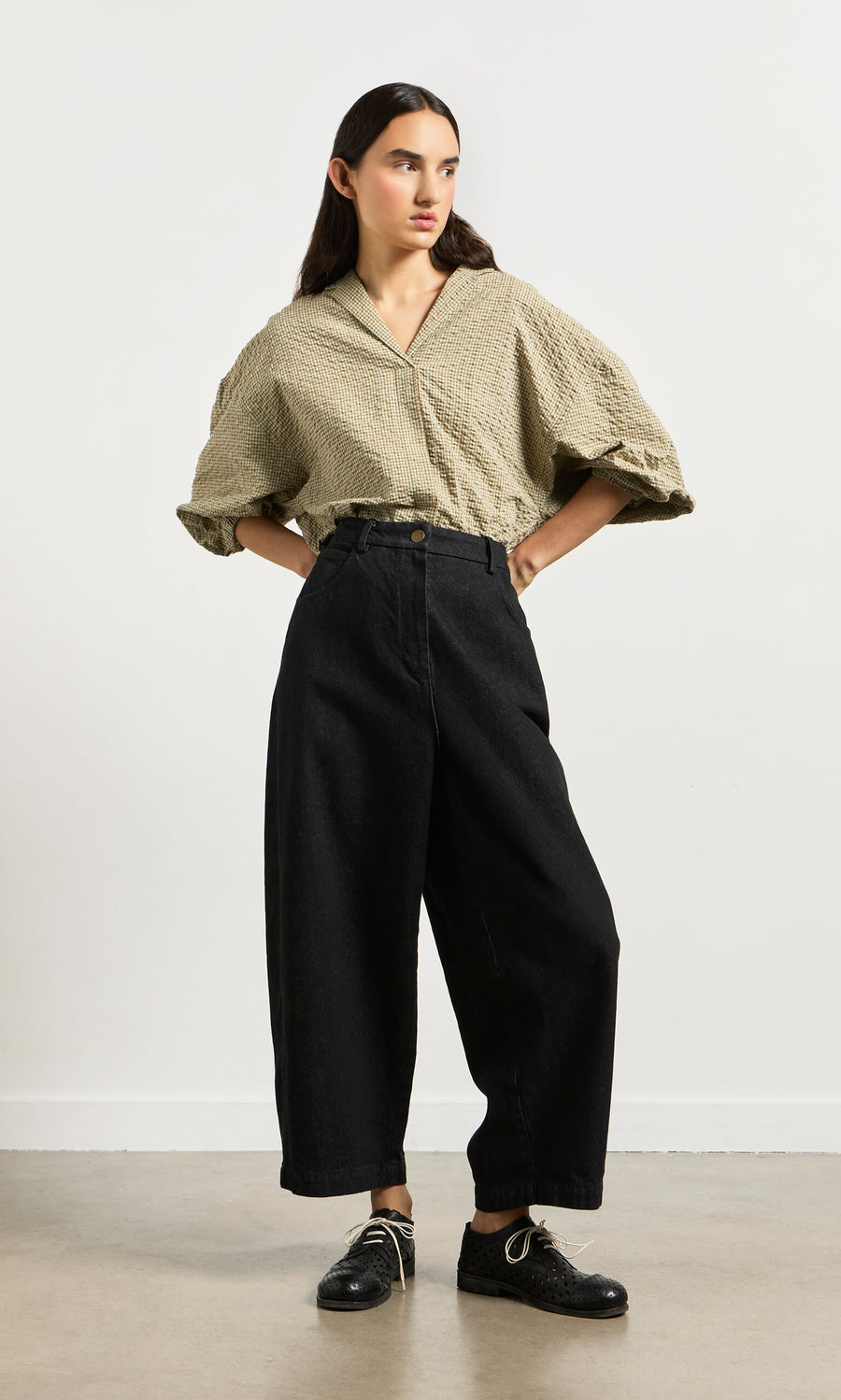 Plümo- Woman stands against a plain wall wearing loose Black Barrel Pants and a beige blouse, embracing a relaxed silhouette.